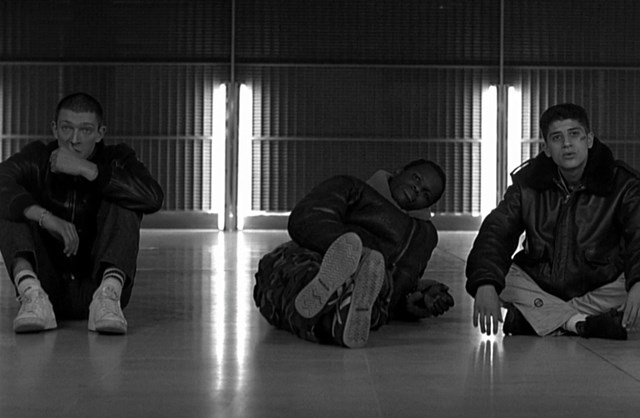 Black-and-white photo of three people sitting and reclining on a smooth floor in front of a wall with vertical panels and bright lights. All are wearing dark jackets and light-colored pants, with casual footwear. The central figure is lying on one side with legs extended, while the others sit cross-legged or with knees bent, creating a relaxed, informal pose in an urban indoor setting.