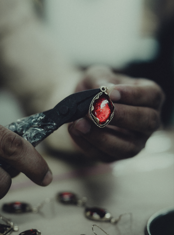 Close-up of artisan hands using pliers to craft a pendant with a deep red gemstone in a decorative metal setting, surrounded by similar pieces on a workbench.