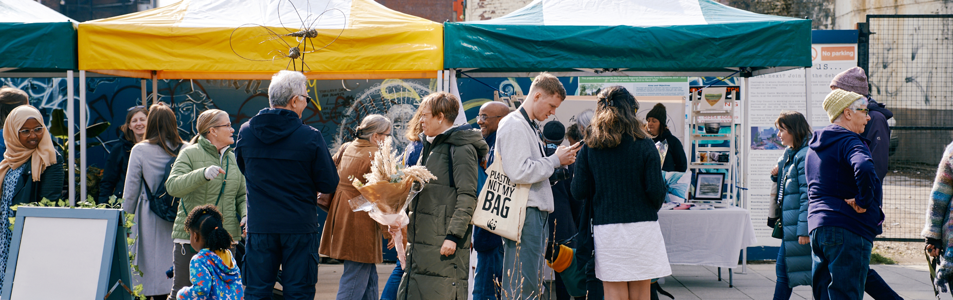 A lively outdoor market scene with people gathered around stalls covered by yellow and green canopies. Various individuals are browsing items, chatting, and holding bags or flowers. Display boards and tables with informational materials are set up under the tents. The background shows older brick buildings and a tall modern structure, while the foreground features plants and a paved area.