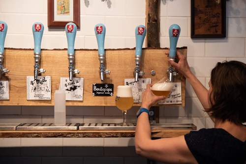 Person pouring a draft beer from a tap in a bar with six blue-handled beer taps mounted on a wooden panel. Two glasses of beer are on the counter, and handwritten labels under the taps display beer names such as ‘Miel’ and ‘Bonpapa.’ White tiled wall in the background.