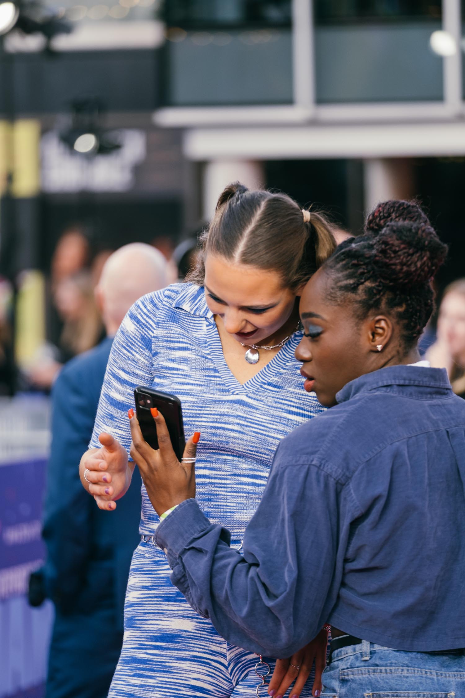Two people standing close together outdoors, one holding a smartphone and showing the screen to the other. The person on the left is wearing a blue patterned outfit with a heart-shaped necklace, while the person on the right is dressed in a dark denim shirt. The background shows a blurred crowd and building structures.