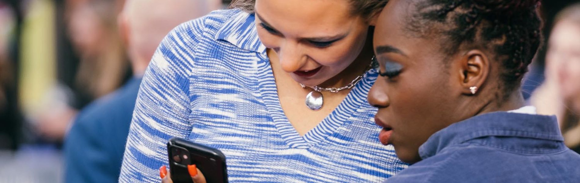 Two people standing close together outdoors, one holding a smartphone and showing the screen to the other. The person on the left is wearing a blue patterned outfit with a heart-shaped necklace, while the person on the right is dressed in a dark denim shirt. The background shows a blurred crowd and building structures.