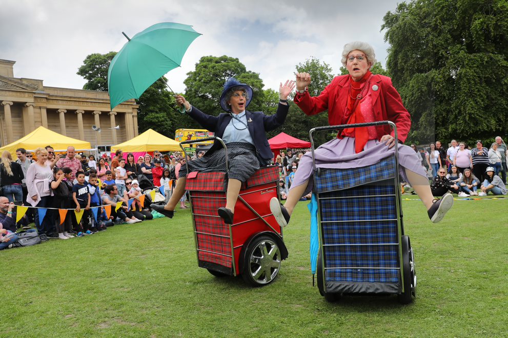 Two performers dressed in vintage-style clothing ride motorised shopping trolleys covered in tartan fabric during an outdoor event. One holds a bright green umbrella while both raise their arms in animated gestures. Behind them, a crowd watches near colourful market-style tents set up on a grassy area with trees and a large classical building in the background.