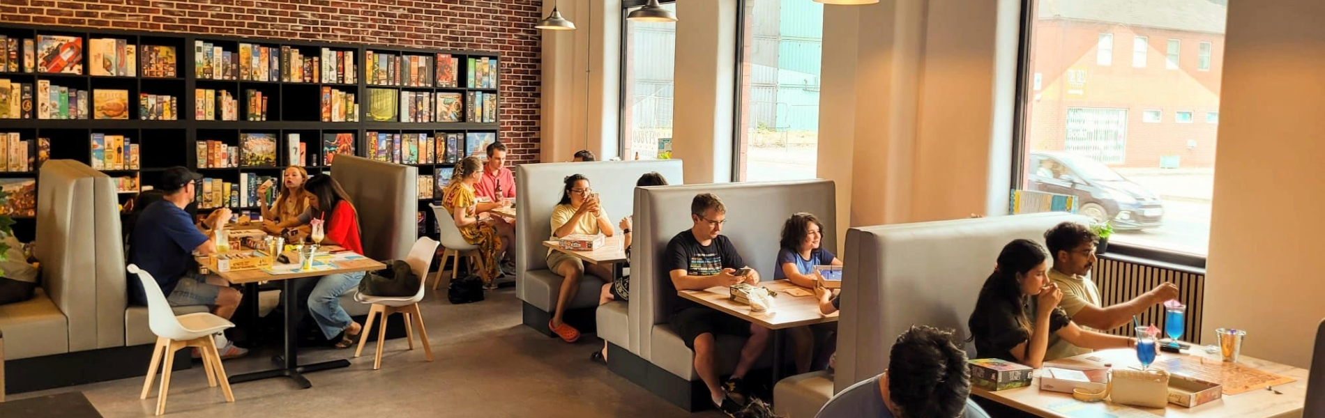 Inside The Dice Box Sheffield, there are tables and booths full of people playing games and drink and eating. The back wall is covered in shelving that is full of boxed games.