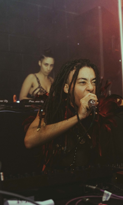 Three performers on stage at No Bounds festival. One is singing into a microphone while the other two are playing records on turntables and using a mixer.