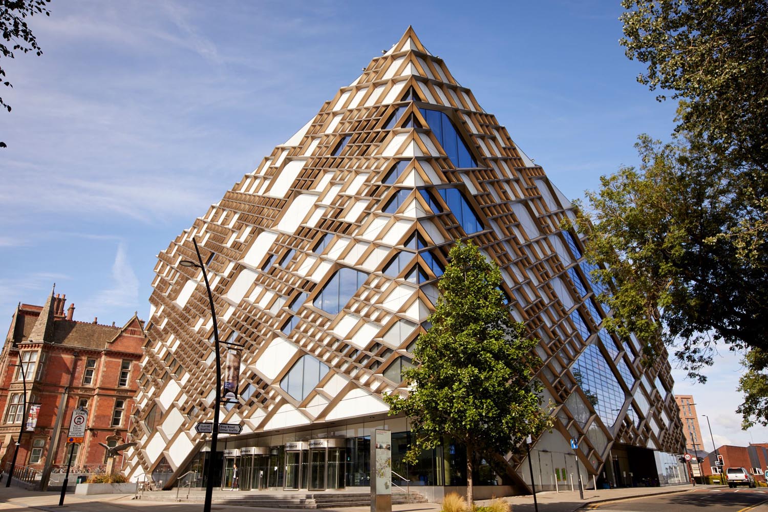 The exterior of the Diamond Building in Sheffield. Striking modern building with a distinctive diamond-shaped design featuring a geometric lattice of wooden beams and large glass panels. The structure has a pyramid-like form with multiple triangular windows, creating a bold architectural statement. Surrounding the building are trees, a paved street, and older brick buildings under a clear blue sky.