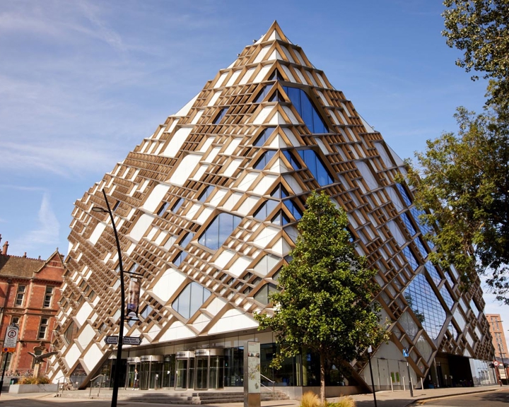 The exterior of the Diamond Building in Sheffield. Striking modern building with a distinctive diamond-shaped design featuring a geometric lattice of wooden beams and large glass panels. The structure has a pyramid-like form with multiple triangular windows, creating a bold architectural statement. Surrounding the building are trees, a paved street, and older brick buildings under a clear blue sky.