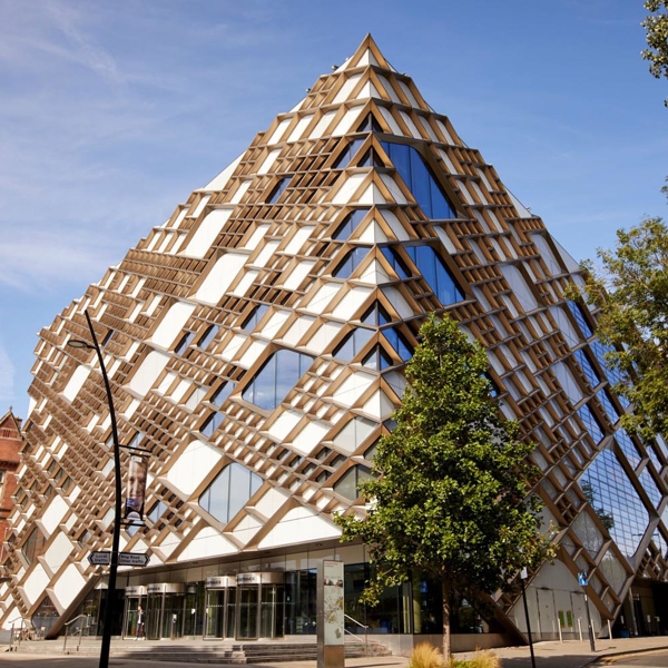 The exterior of the Diamond Building in Sheffield. Striking modern building with a distinctive diamond-shaped design featuring a geometric lattice of wooden beams and large glass panels. The structure has a pyramid-like form with multiple triangular windows, creating a bold architectural statement. Surrounding the building are trees, a paved street, and older brick buildings under a clear blue sky.