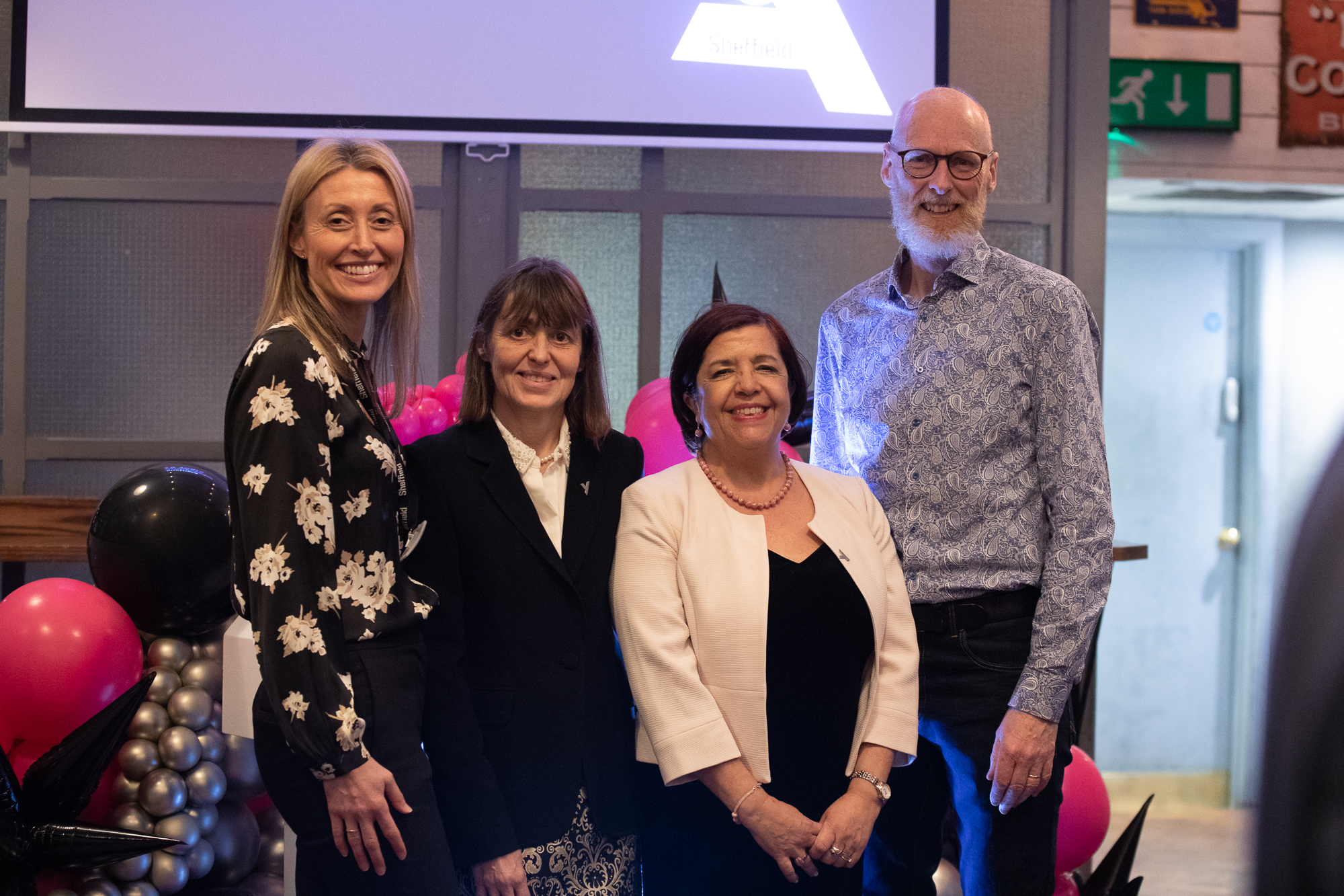 Four people standing together indoors at an event, positioned in front of a backdrop with a large screen displaying a partial logo. The group is surrounded by decorative balloon arrangements in black, pink, and silver. The setting includes grey panelled walls and visible signage in the background, suggesting a formal or celebratory occasion.