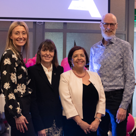 Four people standing together indoors at an event, positioned in front of a backdrop with a large screen displaying a partial logo. The group is surrounded by decorative balloon arrangements in black, pink, and silver. The setting includes grey panelled walls and visible signage in the background, suggesting a formal or celebratory occasion.