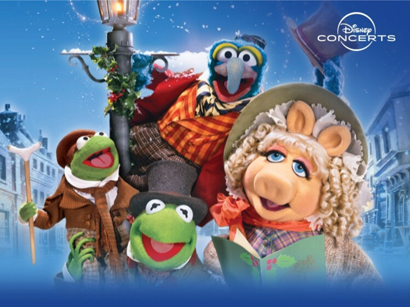 Colorful promotional artwork for Disney Concerts featuring characters from The Muppet Christmas Carol. Kermit the Frog appears twice, dressed in Victorian-style clothing, one holding a walking stick and another wearing a top hat. Gonzo is perched on a lamppost wrapped in festive greenery, wearing a striped scarf and red coat. The background shows a snowy street scene with old-fashioned buildings under a bright blue sky.