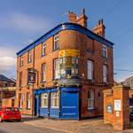 The Fat Cat pub in Kelham Island - Historic red-brick pub building with bright blue trim on a street corner, featuring a large painted sign reading ‘The Victoria Junction Inn.’ A red car is parked nearby, and industrial-style buildings and a tall chimney are visible in the background under a clear blue sky.