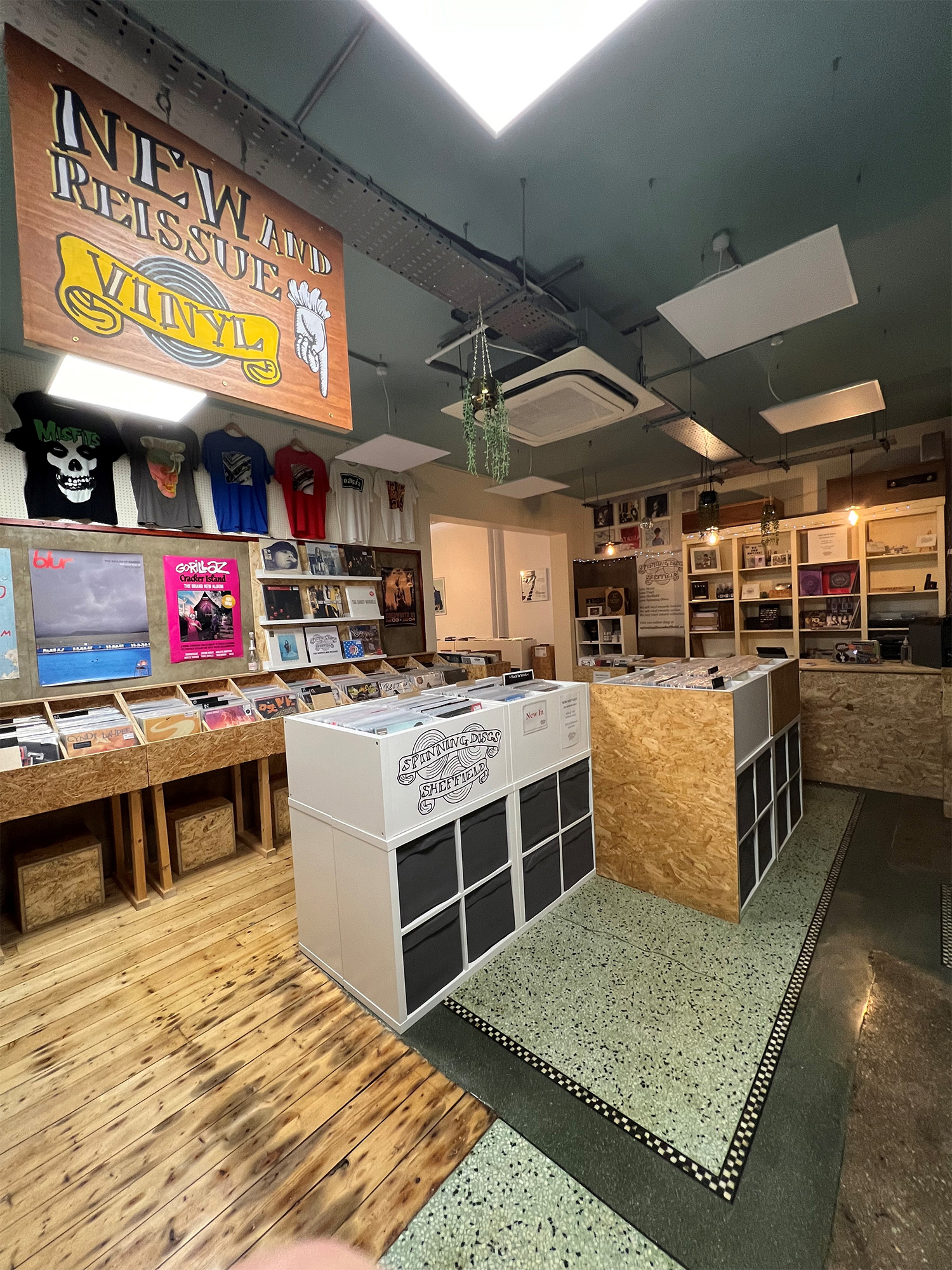 Interior of a record shop featuring wooden and white display units filled with vinyl records. A large sign overhead reads “NEW and REISSUE VINYL.” The walls display colorful posters and T-shirts, and the shop has a mix of wooden and terrazzo flooring. Hanging plants and ceiling lights add to the decor.
