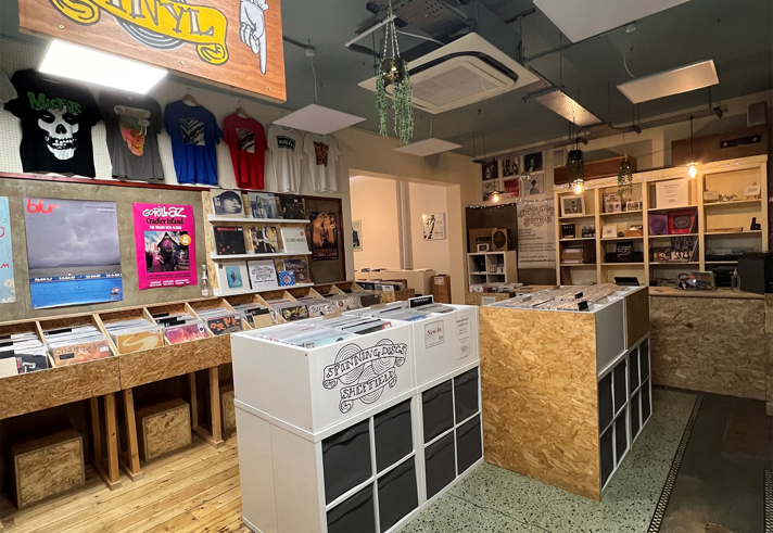 Interior of a record shop featuring wooden and white display units filled with vinyl records. A large sign overhead reads “NEW and REISSUE VINYL.” The walls display colorful posters and T-shirts, and the shop has a mix of wooden and terrazzo flooring. Hanging plants and ceiling lights add to the decor.