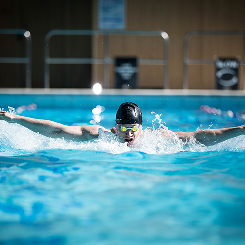 Swimmer performing the butterfly stroke in an indoor pool, arms extended outward and water splashing around. The swimmer is wearing a black swim cap, and the background shows poolside railings and signage.