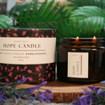 Hope Candle Scent No.7 in Velvet Rose & Oud fragrance, featuring a black and pink floral-patterned box and a lit amber glass jar candle with two wicks, displayed on a wooden base surrounded by green leaves and purple flowers.
