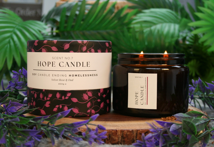 Hope Candle Scent No.7 in Velvet Rose & Oud fragrance, featuring a black and pink floral-patterned box and a lit amber glass jar candle with two wicks, displayed on a wooden base surrounded by green leaves and purple flowers.