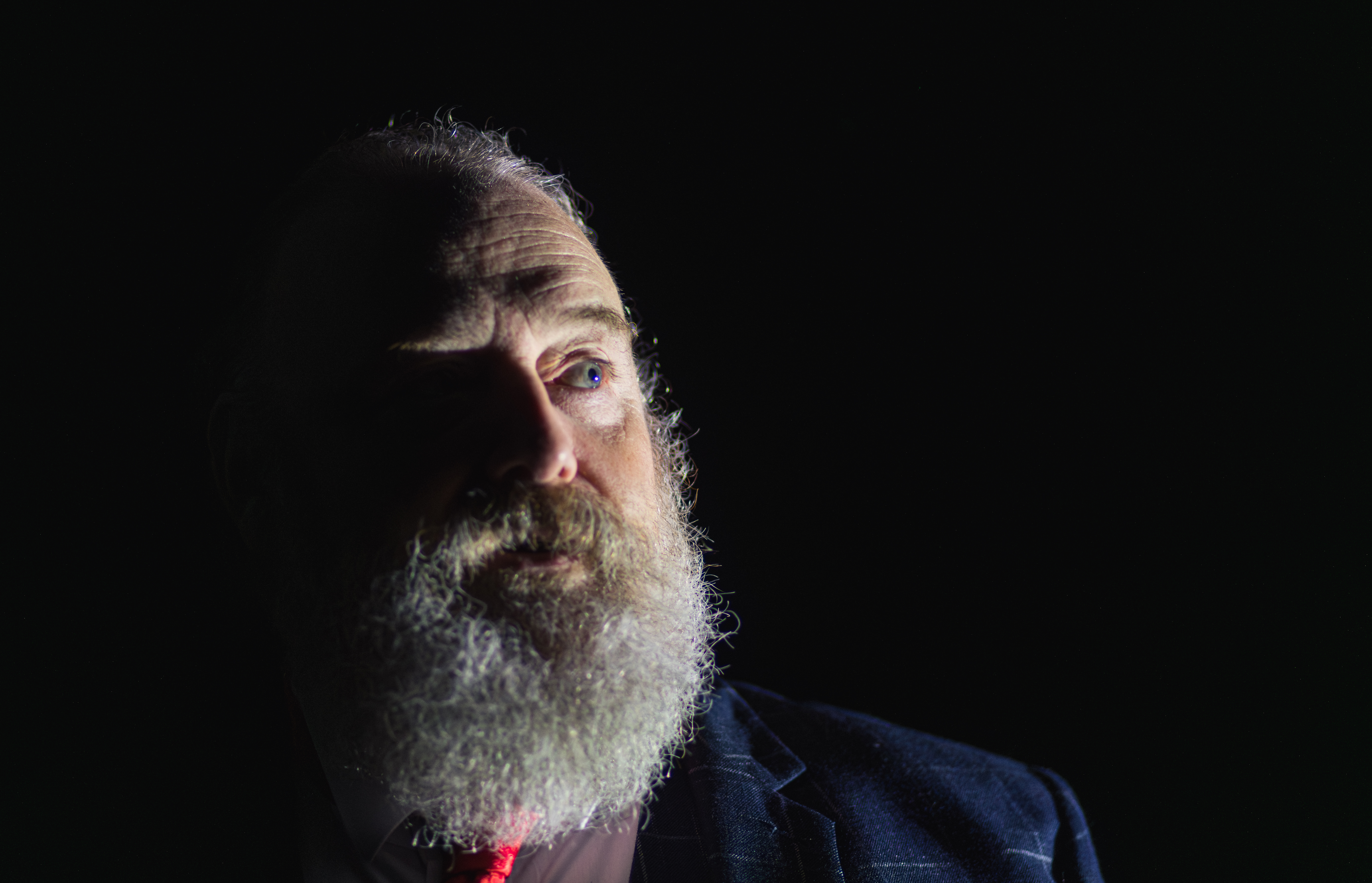 A photograph of a person with a large grey beard standing against a black background. The left side of their face is in shadow.