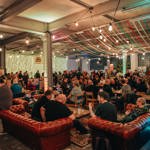 A large indoor festival space filled with people seated at tables and standing in groups. Colourful ribbons hang across the ceiling, and string lights illuminate the room. The atmosphere appears busy and social, with many attendees enjoying drinks.