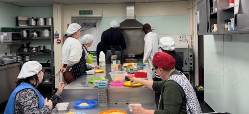 A group of people in a commercial-style kitchen participating in a cooking activity. Several individuals wearing aprons and white hairnets are gathered around a stainless steel worktable with colorful cutting boards, bowls, and various ingredients. Some are seated while others stand near a large stove and extractor hood at the back of the room. Shelves with kitchen equipment and containers line the walls, and the overall setting appears organized and functional for group cooking.