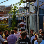 A singer performing with a guitar on a small outdoor stage, surrounded by metal frames and plants. People sit at picnic tables listening and drinking in a relaxed, sunny setting.