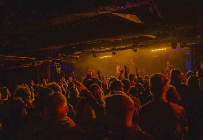 A packed audience inside a low‑lit venue faces a stage with bright amber lighting, where musicians perform. Raised hands and silhouettes fill the scene, creating a lively concert atmosphere.