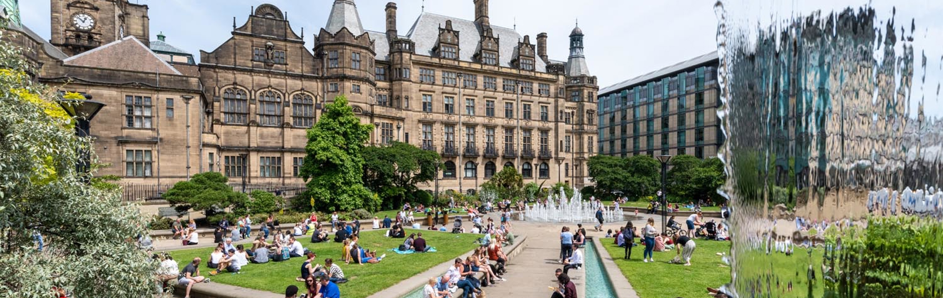 The Peace Gardens, in the centre of Sheffield, on a sunny day. There are lots of people sat on the grass and on benches enjoying the sunshine. In the background is Sheffield Town Hall.