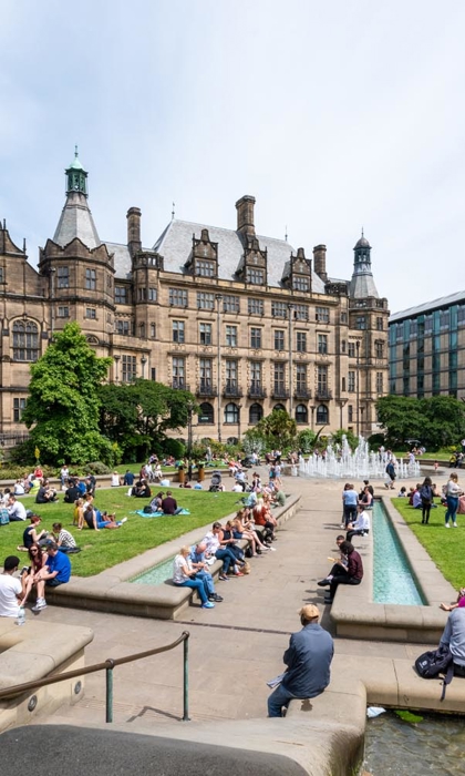 The Peace Gardens, in the centre of Sheffield, on a sunny day. There are lots of people sat on the grass and on benches enjoying the sunshine. In the background is Sheffield Town Hall.