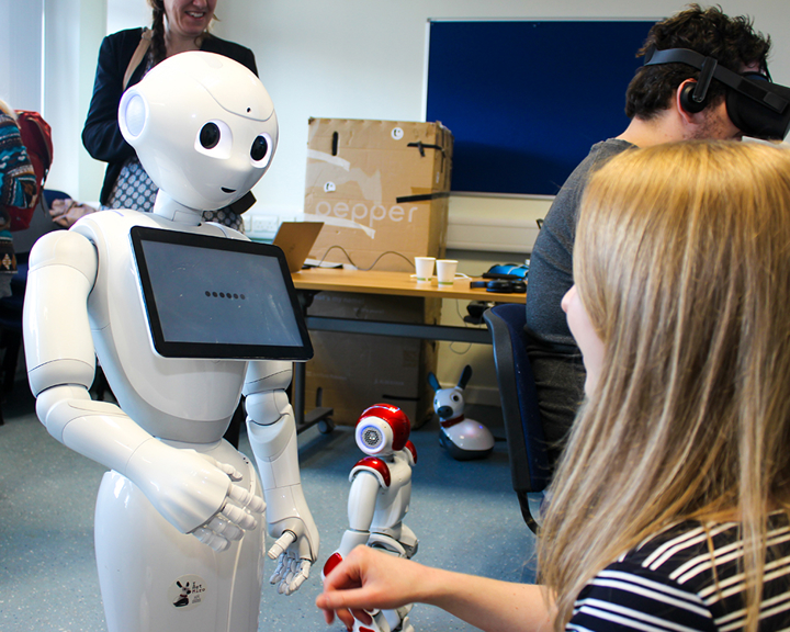 A woman interacting with a humanoid robot which has a touch screen on its chest. There are three other people in the room, one of whom is wearing a virtual reality headset.