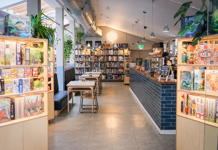 Inside The Treehouse Cafe. You can see tables and chairs, the counter and three shelving units full of board games.