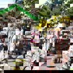  An outdoor market stall displays a variety of colourful wool blankets, throws, and ponchos arranged on racks, mannequins, and benches. A green canopy shades the stall, which also features cushions and a round pouffe in matching patterns. A wooden sign reads “Yorkshire Blankets” alongside smaller signs for luxury wool items. Bright yellow flowers and lush greenery surround the area, creating a vibrant and inviting setting.