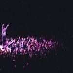 A performer stands on an elevated platform in front of a densely packed crowd, with hundreds of raised hands reaching toward the stage. The scene is lit with deep pink and purple lighting against a dark background.