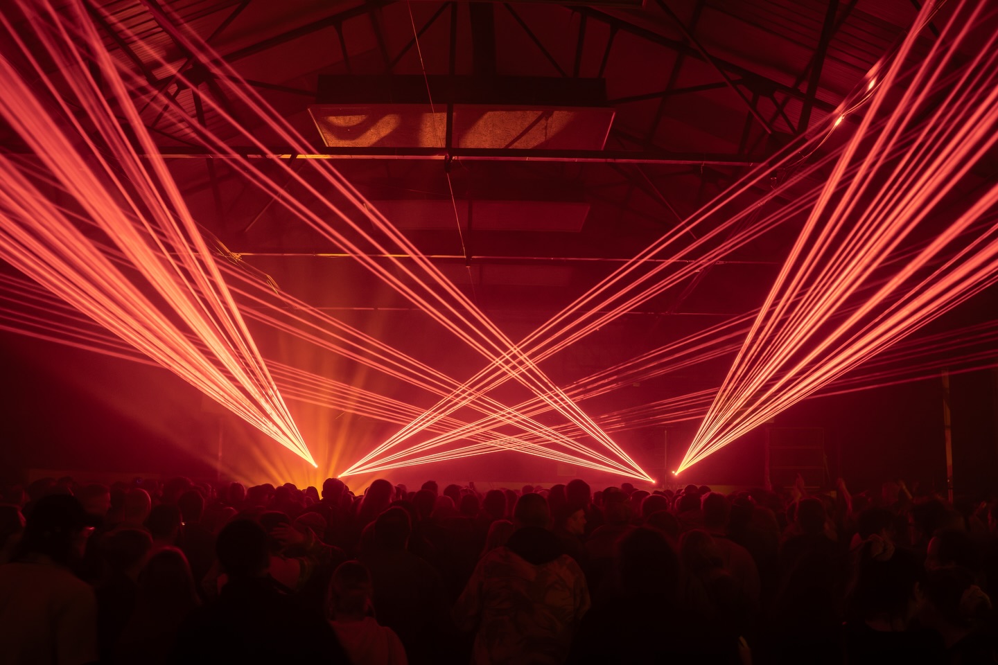 A large indoor crowd watching an intense light show as multiple red laser beams fan out across a dark warehouse-style venue with high, exposed rafters.
