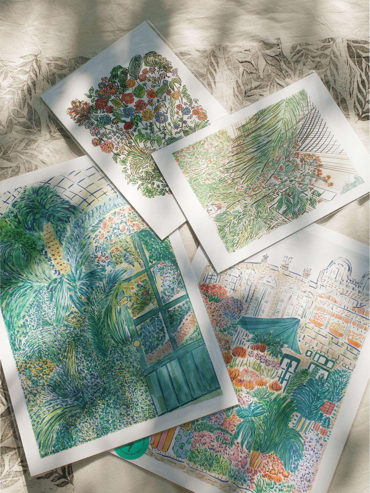 Four botanical art prints arranged on a light fabric surface with dappled sunlight. The prints depict lush greenery, tropical plants, and floral compositions in vivid colors, including one featuring a garden scene with a door and another showing rooftops and foliage.