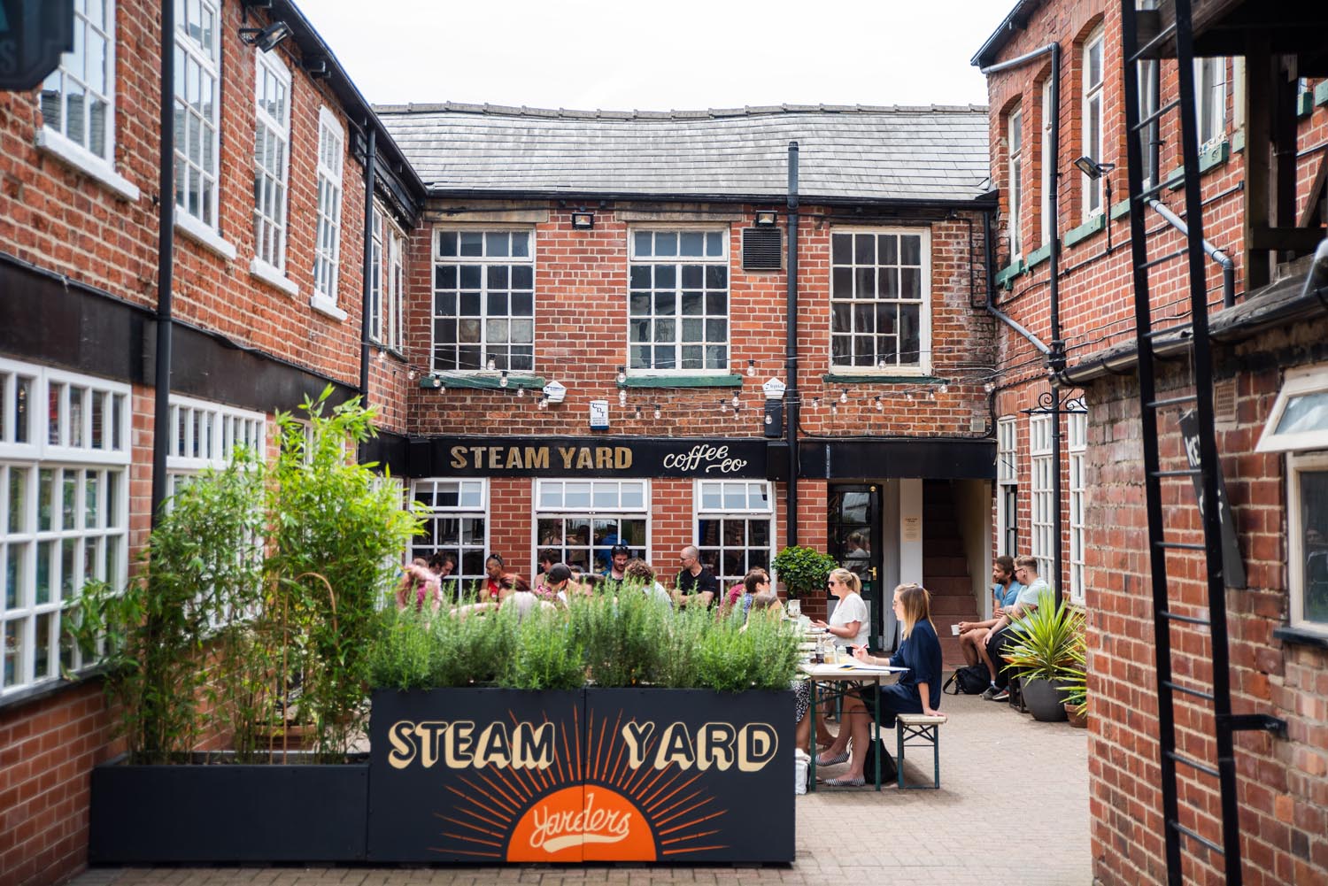 Outdoor seating area at Steam Yard coffee shop in a red-brick courtyard. Several people are sitting at tables enjoying drinks. The shop’s black sign reads “Steam Yard coffee co.” Large planter with green plants in the foreground displays “Steam Yard” and “Javelons” in bold lettering. Surrounding buildings have white-framed windows and a rustic industrial look.