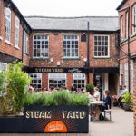 Outdoor seating area at Steam Yard coffee shop in a red-brick courtyard. Several people are sitting at tables enjoying drinks. The shop’s black sign reads “Steam Yard coffee co.” Large planter with green plants in the foreground displays “Steam Yard” and “Javelons” in bold lettering. Surrounding buildings have white-framed windows and a rustic industrial look.