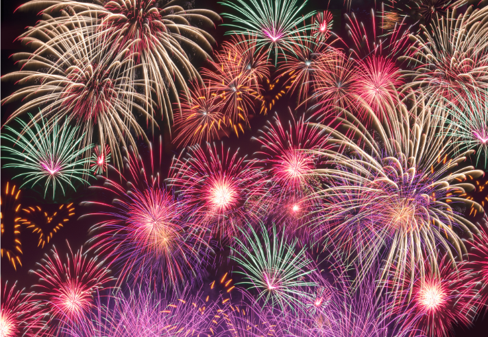 A vibrant display of fireworks bursting against a dark night sky. Multiple colorful explosions in shades of red, pink, purple, green, and gold fill the scene, creating overlapping patterns of light and streaks. The fireworks vary in size and shape, with some forming large circular bursts and others radiating fine trails, giving a festive and celebratory atmosphere.
