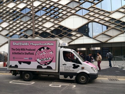 A delivery truck parked on a city street in front of a modern building with a geometric glass facade. The truck has a bright pink advertisement on its side that reads: “Sheffield’s #SuperFreshMilk – The Only Milk Produced & Bottled in Sheffield” along with the logo “Our Cow Molly Dairy Farm.” The design includes black cow-print patches and social media icons. Several people are walking on the pavement near metal barriers in the background.