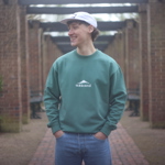 Person wearing a forest green sweatshirt with the “Sabi & Soul” logo printed in white on the chest, paired with light blue jeans and a white cap. The setting is an outdoor walkway framed by brick pillars and wooden beams.