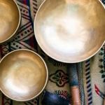 A set of metal singing bowls of varying sizes is arranged on a patterned woven fabric with geometric designs. Two wooden mallets with padded ends lie beside the bowls. The scene is viewed from above, showing the smooth, reflective surfaces of the bowls and the detailed textile beneath them.