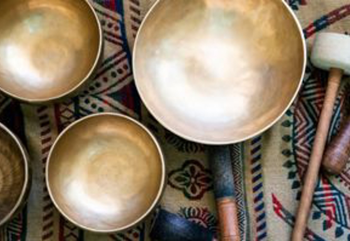 A set of metal singing bowls of varying sizes is arranged on a patterned woven fabric with geometric designs. Two wooden mallets with padded ends lie beside the bowls. The scene is viewed from above, showing the smooth, reflective surfaces of the bowls and the detailed textile beneath them.