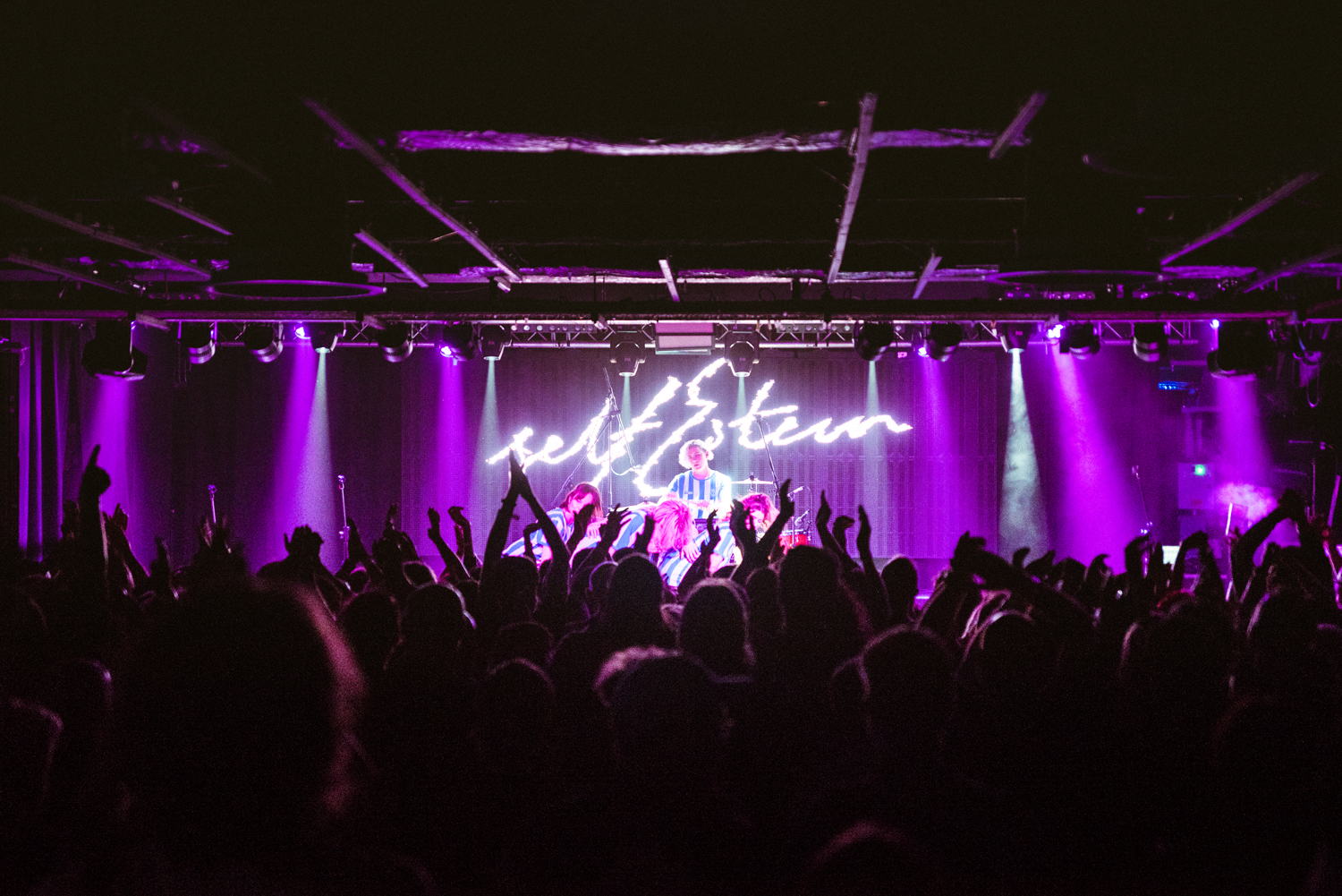 A crowded concert venue with an audience raising hands and cheering in front of a brightly lit stage. The stage is illuminated with vibrant purple lighting, and a performer stands at the center under a glowing neon-style sign that reads “self esteem.” The ceiling has exposed beams, and the atmosphere suggests an energetic live music event.