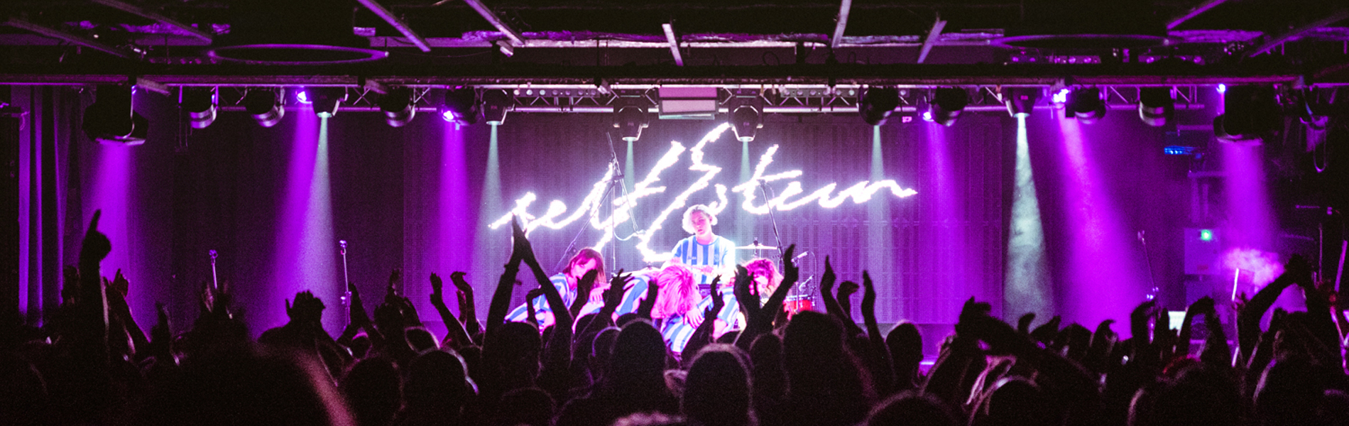 A crowded concert venue with an audience raising hands and cheering in front of a brightly lit stage. The stage is illuminated with vibrant purple lighting, and a performer stands at the center under a glowing neon-style sign that reads “self esteem.” The ceiling has exposed beams, and the atmosphere suggests an energetic live music event.