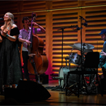 A band performs on a stage, with a woman singing and two men playing upright bass and drums.