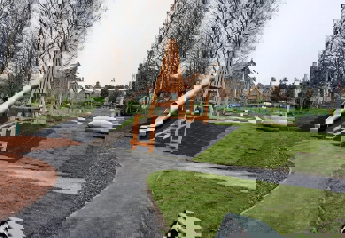 Playground with a large wooden-framed structure and a long, curved metal slide. The area is surrounded by grass, pathways, and leafless trees. Stone buildings and fencing are visible in the background under an overcast sky.