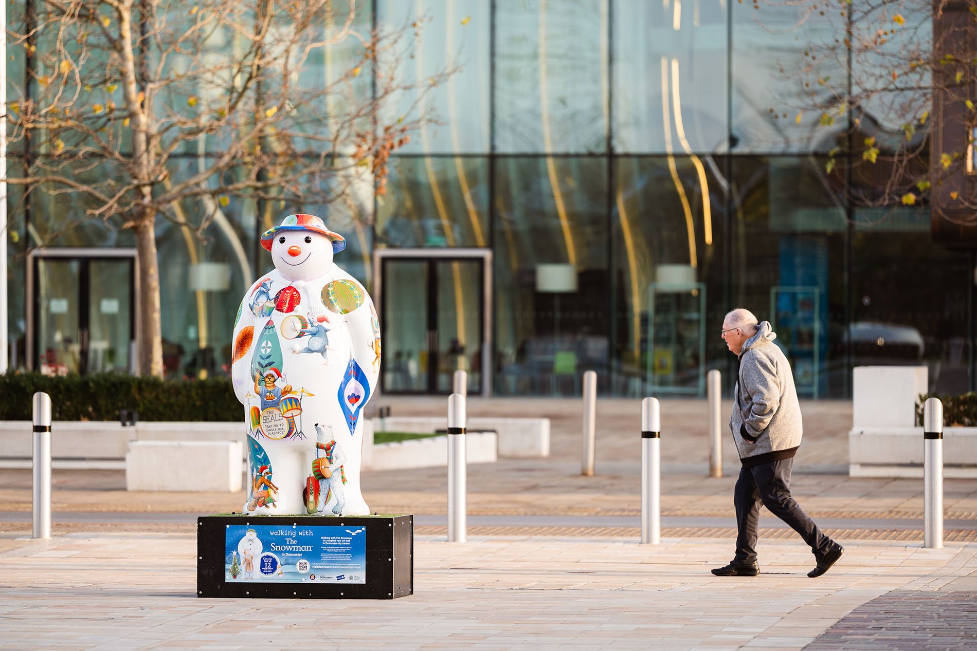 A white snowman sculpture decorated with colorful illustrations, including festive designs and patterns, stands on a black platform in a paved outdoor area. Behind the sculpture is a modern glass building with tall windows and reflections, and leafless trees line the space.