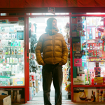A man in a yellow puffa jacket stands outside a convenience store at night.