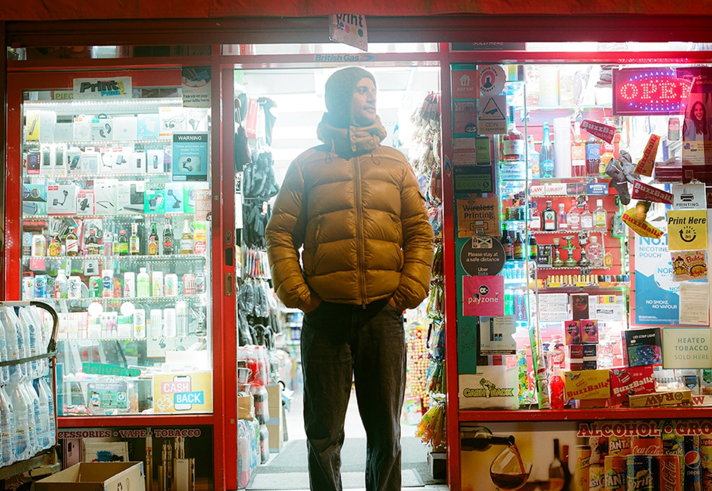 A man in a yellow puffa jacket stands outside a convenience store at night.