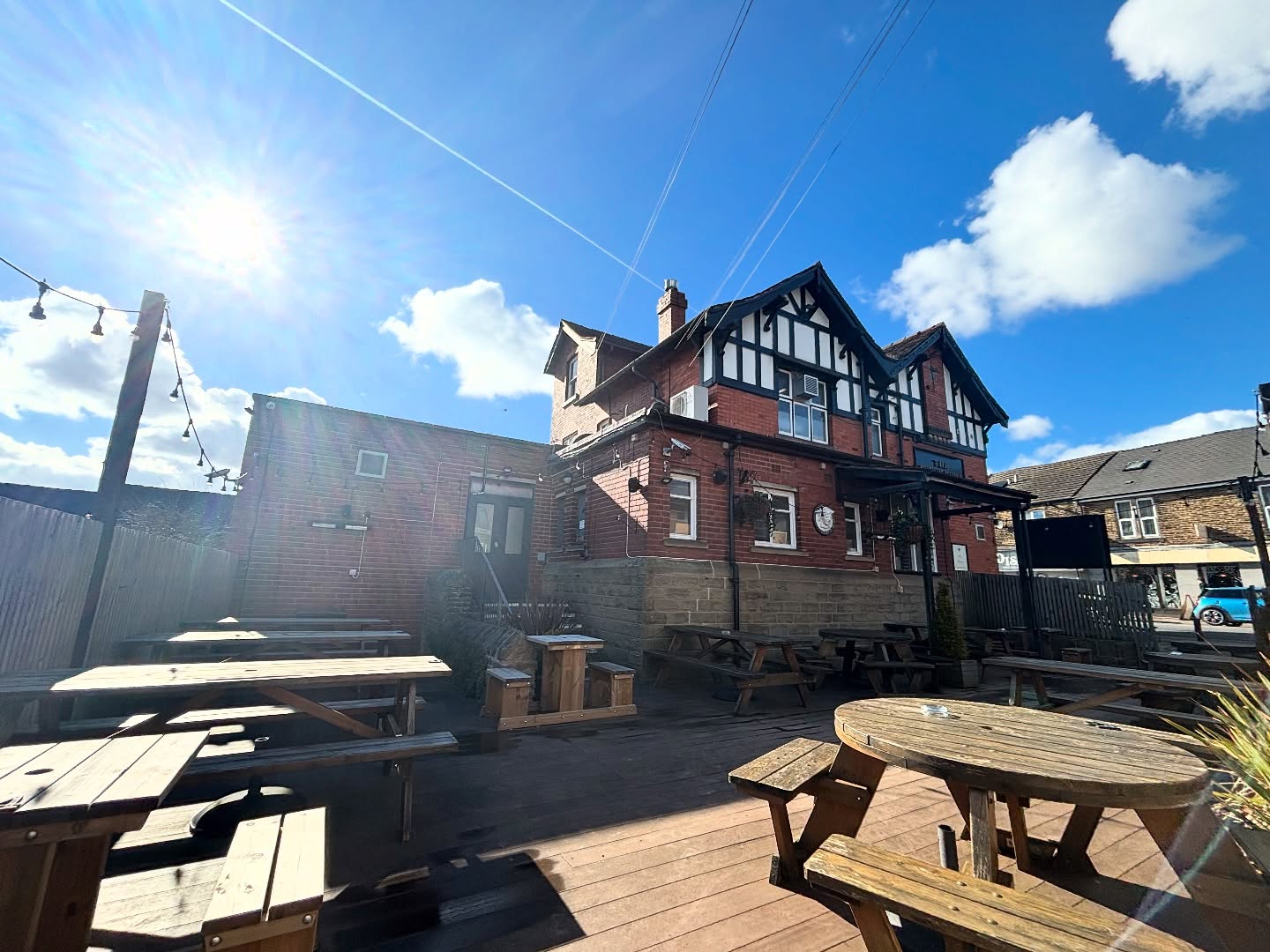 A sunlit outdoor decking area behind The Punch Bowl, with wooden picnic tables and string lights, and the pub building visible in the background.