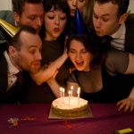 A group of people in party hats crowd round a birthday, blowing out the candles.